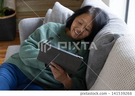 Senior asian woman reclining on couch beside woven planter basket holding tablet computer Senior asian woman reclining on couch beside woven planter basket holding tablet computer 141539268