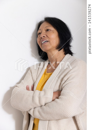 Senior Asian woman leaning against white wall in studio wearing mustard shirt and beige cardigan Senior Asian woman leaning against white wall in studio wearing mustard shirt and beige cardigan 141539274