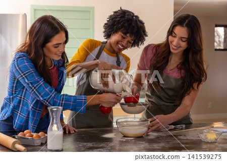 Diverse female friends measuring flour using cup and whisking batter in bowl at kitchen island Diverse female friends measuring flour using cup and whisking batter in bowl at kitchen island 141539275