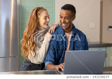 Diverse couple leaning on stone countertop in kitchen next to green cabinets while using laptop Diverse couple leaning on stone countertop in kitchen next to green cabinets while using laptop 141539277