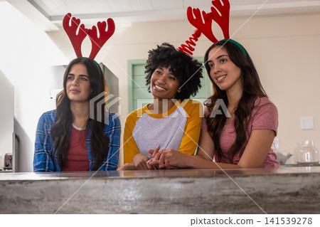 Diverse female friends in antler headbands sitting at stone kitchen counter near glass jar Diverse female friends in antler headbands sitting at stone kitchen counter near glass jar 141539278