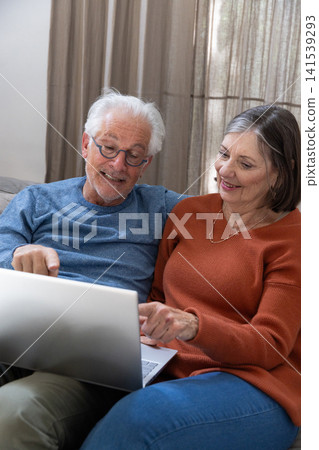Senior couple leaning forward toward silver laptop on neutral-tone sofa in home living room Senior couple leaning forward toward silver laptop on neutral-tone sofa in home living room 141539293