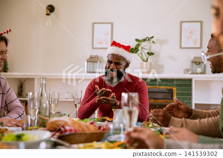 Diverse friends laughing, clapping at dining table at home with champagne flutes, santa hat Diverse friends laughing, clapping at dining table at home with champagne flutes, santa hat 141539302
