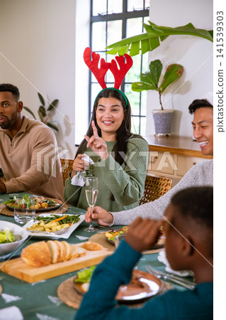Diverse family sitting at home dining table with woman in antlers holding napkin by salad bowl Diverse family sitting at home dining table with woman in antlers holding napkin by salad bowl 141539303