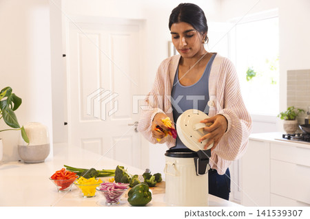 Asian woman placing chopped vegetables into white slow cooker on kitchen island at home 141539307