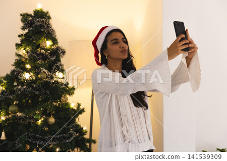 Indian woman wearing Santa hat taking selfie with smartphone in living room by Christmas tree 141539309