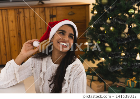 South Asian woman sitting at home wearing sweater and red Santa hat by Christmas tree smiling South Asian woman sitting at home wearing sweater and red Santa hat by Christmas tree smiling 141539312