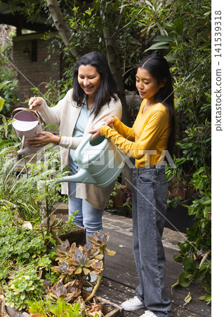 Diverse mother and daughter watering plants on deck in backyard with pink and green watering cans 141539318