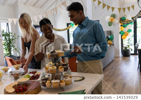 Diverse friends tasting cupcakes and chips around dining table at home with balloons, banners Diverse friends tasting cupcakes and chips around dining table at home with balloons, banners 141539329