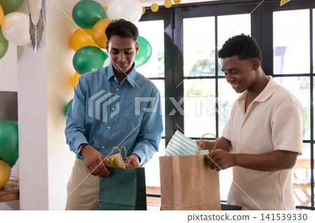 Diverse male friends standing at table opening gift bags amid balloon banner display Diverse male friends standing at table opening gift bags amid balloon banner display 141539330
