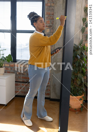 African American woman wearing headphones writing on board using yellow marker and tablet at office African American woman wearing headphones writing on board using yellow marker and tablet at office 141539341