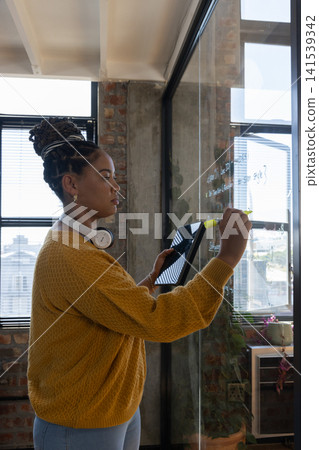 African American woman writing notes on glass board in loft office holding tablet, copy space African American woman writing notes on glass board in loft office holding tablet, copy space 141539342