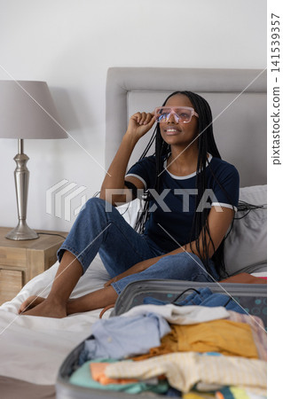 African American woman packing suitcase on bed in bedroom adjusting pink eyeglasses, copy space African American woman packing suitcase on bed in bedroom adjusting pink eyeglasses, copy space 141539357