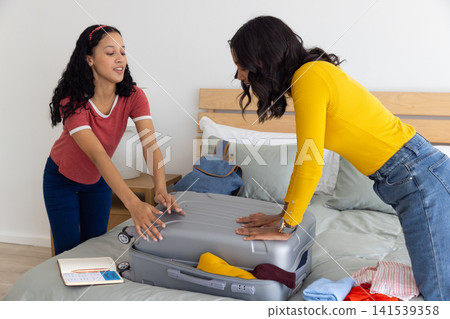 Diverse mom and teenage daughter packing clothes in suitcase on bed with notebook and backpack Diverse mom and teenage daughter packing clothes in suitcase on bed with notebook and backpack 141539358
