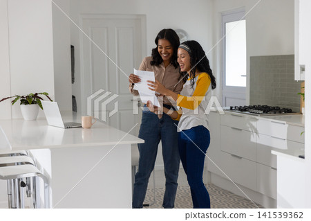Diverse mom and teenage daughter reviewing papers at white island with laptop and mug Diverse mom and teenage daughter reviewing papers at white island with laptop and mug 141539362