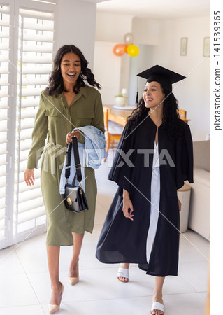 Diverse mother with teenage daughter in living room wearing gown and mortarboard cap Diverse mother with teenage daughter in living room wearing gown and mortarboard cap 141539365