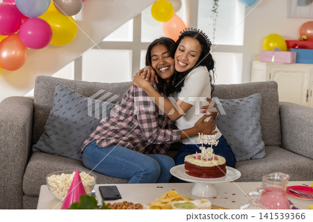 Diverse mom and teenage daughter hugging on grey couch in living room near birthday cake candles Diverse mom and teenage daughter hugging on grey couch in living room near birthday cake candles 141539366