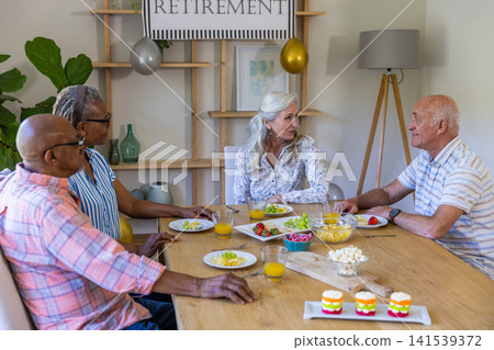 Diverse senior friends chatting at home dining table with fruit, juice under retirement banner 141539372