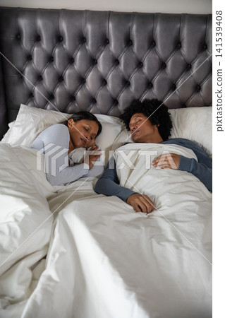 Lying under white duvet on bed with tufted headboard, diverse couple in bedroom Lying under white duvet on bed with tufted headboard, diverse couple in bedroom 141539408