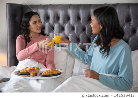 Diverse female friends sitting up in bed and clinking glasses of orange juice over croissants Diverse female friends sitting up in bed and clinking glasses of orange juice over croissants 141539412