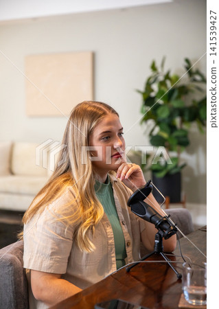 Female content creator recording podcast at table in living room with microphone, glass of water Female content creator recording podcast at table in living room with microphone, glass of water 141539427