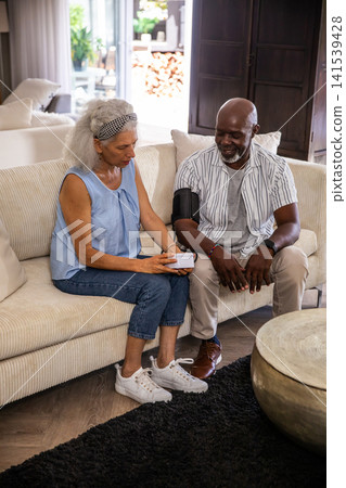 Senior couple measuring blood pressure with portable monitor sitting on cream sofa in living room Senior couple measuring blood pressure with portable monitor sitting on cream sofa in living room 141539428