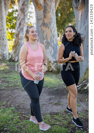 Diverse female friends walking along dirt path in park holding water bottles, wearing smartwatch Diverse female friends walking along dirt path in park holding water bottles, wearing smartwatch 141539429