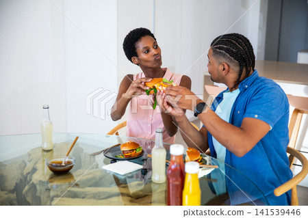 Diverse couple holding hamburgers leaning at table in kitchen with drink bottles and sauce bowl Diverse couple holding hamburgers leaning at table in kitchen with drink bottles and sauce bowl 141539446