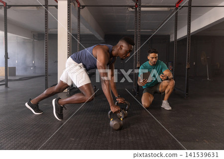 Male african american trainee, trainer guiding kettlebell climbers at gym with tablet, stopwatch Male african american trainee, trainer guiding kettlebell climbers at gym with tablet, stopwatch 141539631