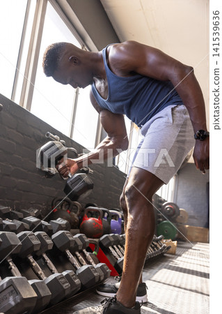 African american man wearing sportswear lifting dumbbell by kettlebells at weight room African american man wearing sportswear lifting dumbbell by kettlebells at weight room 141539636