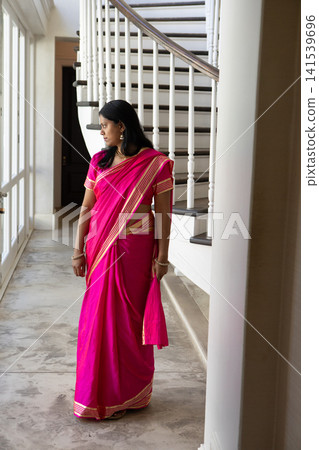 Indian woman standing in foyer, wearing vibrant pink saree and gold jewelry by tall windows Indian woman standing in foyer, wearing vibrant pink saree and gold jewelry by tall windows 141539696