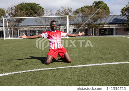 African American man wearing jersey kneeling on turf field at soccer goal cheering with arms raised African American man wearing jersey kneeling on turf field at soccer goal cheering with arms raised 141539713