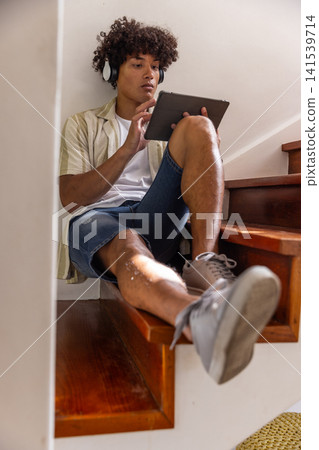African american man sitting on staircase at home using tablet with wireless headphones African american man sitting on staircase at home using tablet with wireless headphones 141539714