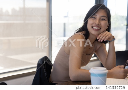 Asian woman leaning at table by window with smartphone beside lidded coffee cup, copy space 141539729
