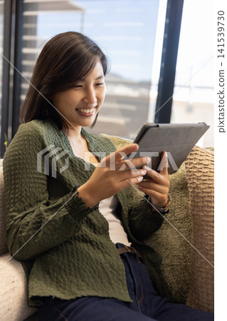 Asian woman sitting on textured sofa in lounge holding tablet with case near decorative cushion Asian woman sitting on textured sofa in lounge holding tablet with case near decorative cushion 141539730