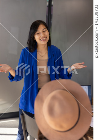 Korean woman in blazer presenting and gesturing at meeting desk with laptop, papers, brown hat Korean woman in blazer presenting and gesturing at meeting desk with laptop, papers, brown hat 141539733