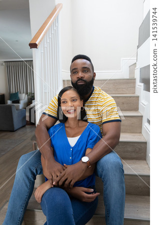 Diverse couple sitting on wooden staircase at home, wrapping arms around and showing wristwatch Diverse couple sitting on wooden staircase at home, wrapping arms around and showing wristwatch 141539744