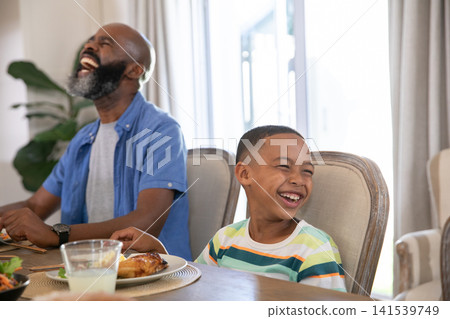 African American father and son laughing at wooden dining table with chicken drumstick and milk African American father and son laughing at wooden dining table with chicken drumstick and milk 141539749