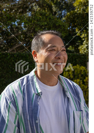 Mature adult asian man standing in sunlit garden wearing striped shirt and t-shirt Mature adult asian man standing in sunlit garden wearing striped shirt and t-shirt 141539752
