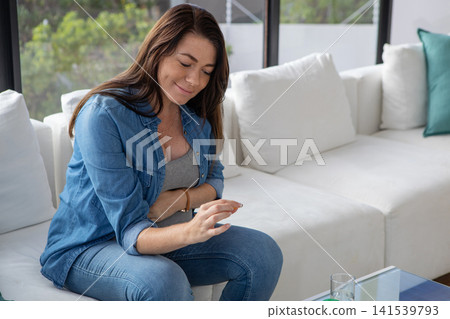 woman sitting on sofa by glass coffee table in living room checking gold bracelet, copy space woman sitting on sofa by glass coffee table in living room checking gold bracelet, copy space 141539793