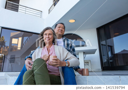 Senior couple sitting on steps under balcony at home holding mugs and gazing thoughtfully 141539794