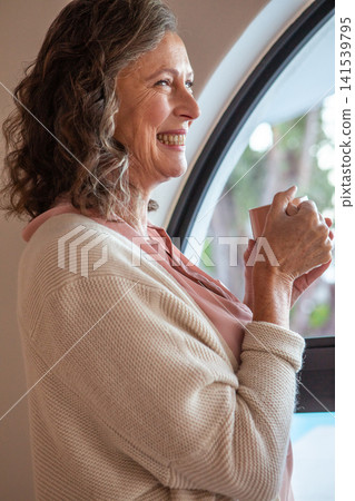 Senior woman standing near arched window at home holding mug wearing cardigan, copy space Senior woman standing near arched window at home holding mug wearing cardigan, copy space 141539795