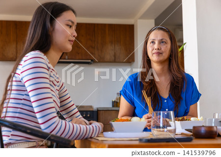 Asian mother and daughter sitting at dining table in kitchen holding chopsticks over bowl Asian mother and daughter sitting at dining table in kitchen holding chopsticks over bowl 141539796