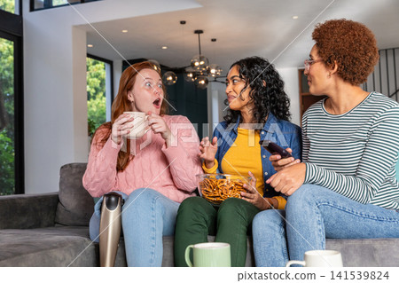 Diverse female friends sitting on living room sofa holding mugs, bowl of pretzels, smartphone Diverse female friends sitting on living room sofa holding mugs, bowl of pretzels, smartphone 141539824