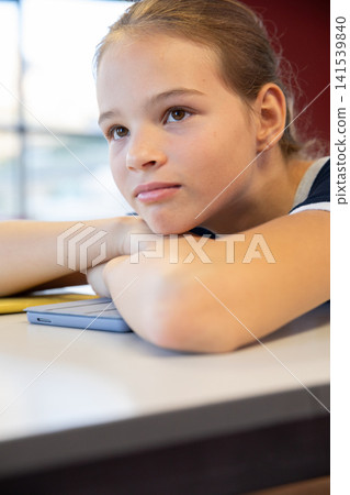Child girl resting head on folded arms at desk in classroom next to bright window with tablet Child girl resting head on folded arms at desk in classroom next to bright window with tablet 141539840