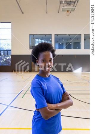 African american teenage athlete standing at court in gym under basketball hoop African american teenage athlete standing at court in gym under basketball hoop 141539841