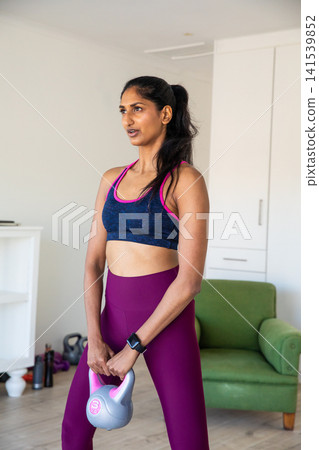 Indian woman performing kettlebell exercise at home holding grey pink kettlebell Indian woman performing kettlebell exercise at home holding grey pink kettlebell 141539852