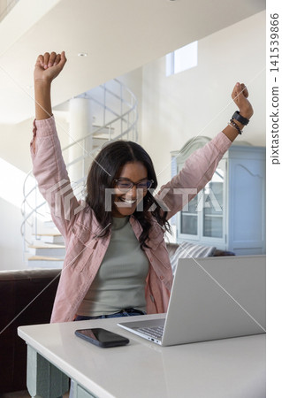African american woman in 20s celebrating at home desk with laptop smartphone bracelets 141539866