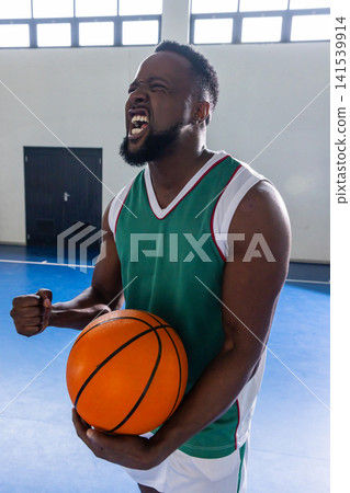African american man holding orange basketball on court at gym in green jersey, white shorts African american man holding orange basketball on court at gym in green jersey, white shorts 141539914