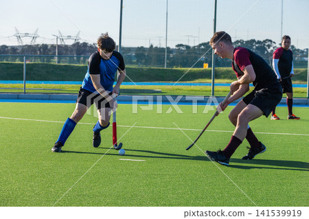 Male field hockey opponents competing for ball on artificial turf pitch with sticks Male field hockey opponents competing for ball on artificial turf pitch with sticks 141539919
